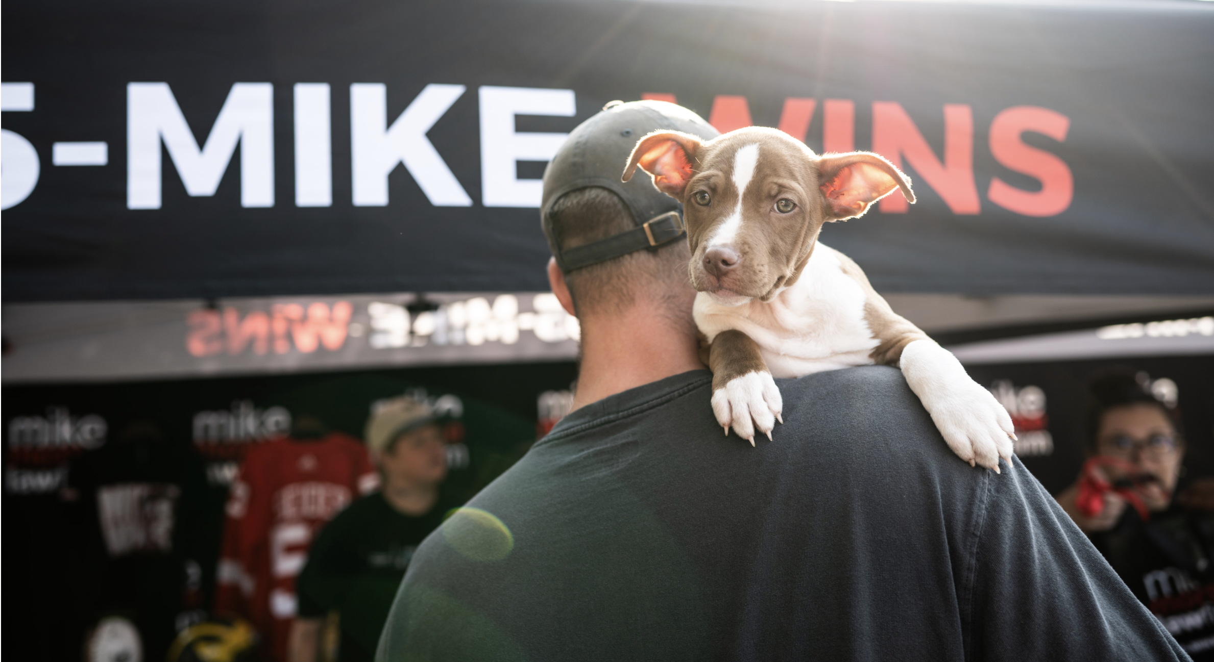 Image of a person wearing a hat. They are faced backwards with a dog over their shoulder facing the camera